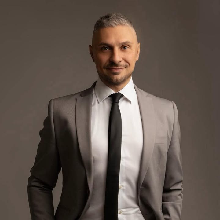 Mauro Mascolo in giacca grigia, camicia bianca e cravatta nera, su fondo grigio